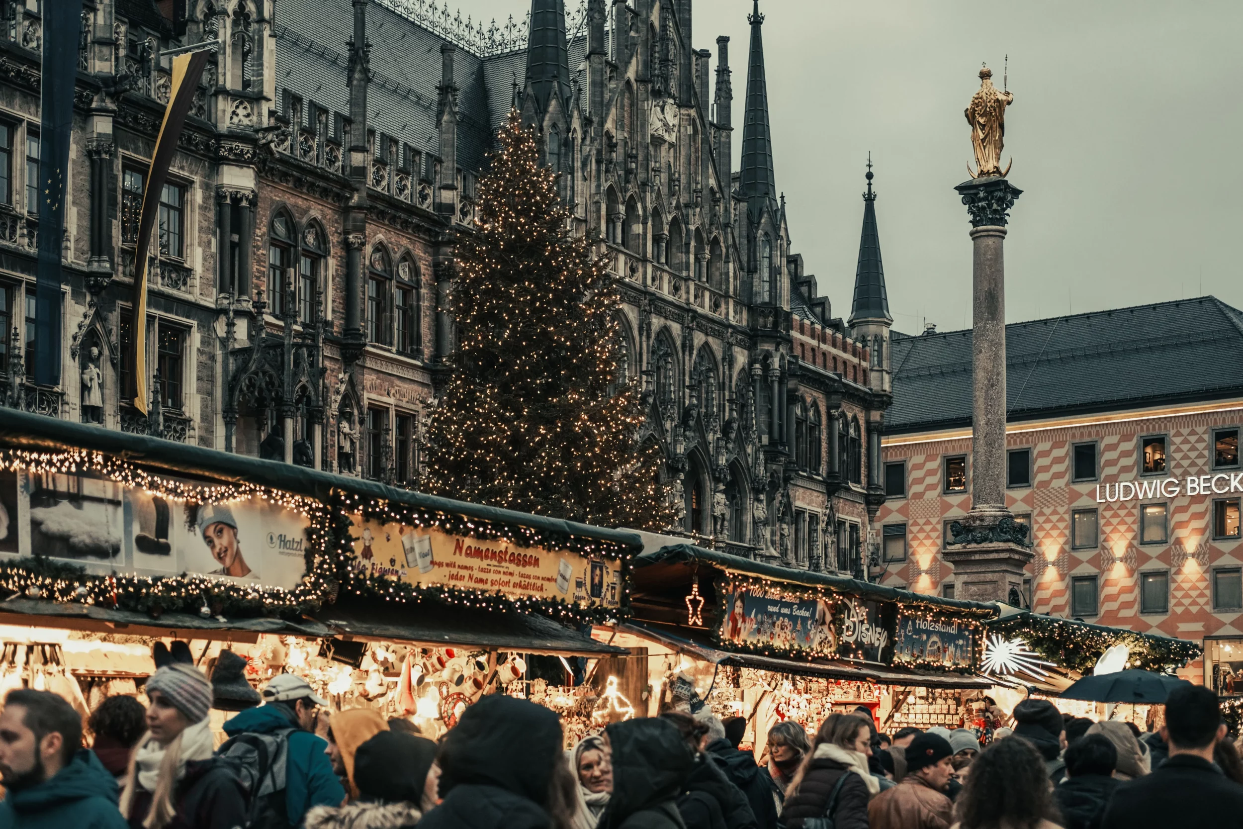 Blick auf den belebten Münchner Christkindlmarkt am Marienplatz am späten Nachmittag, mit festlich erleuchteten Holzbuden, zahlreichen Besuchern und einem großen funkelnden Weihnachtsbaum vor der historischen Kulisse des Neuen Rathauses und der Mariensäule.