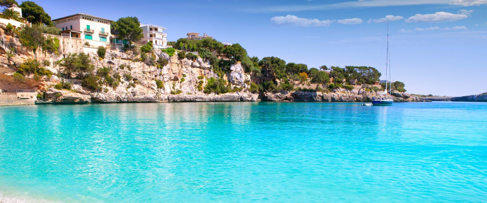 Blick auf eine Bucht mit kristallklarem, türkisfarbenem Wasser in Mallorca, mit einer Felsküste auf der linken Seite und einem Segelboot im Wasser.