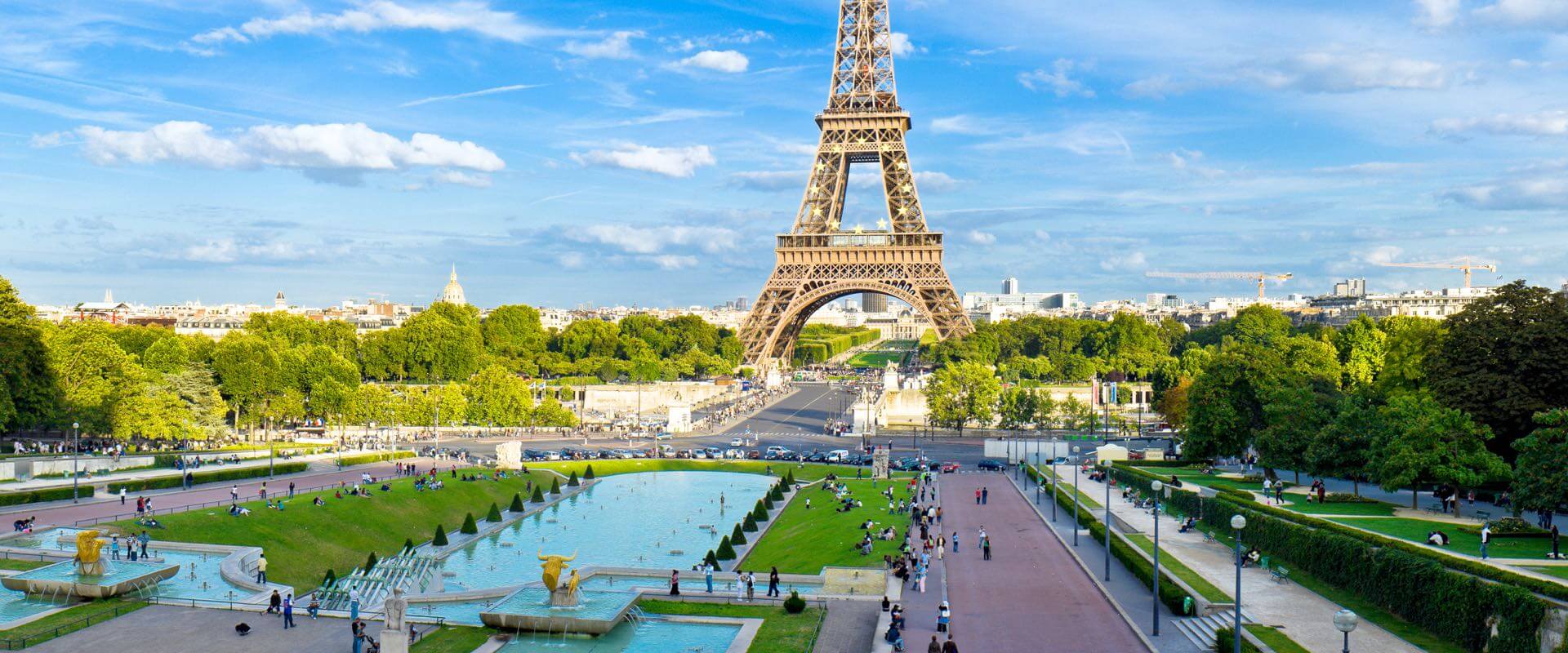 Blick auf den Eiffelturm in Paris, mit den Trocadéro-Gärten im Vordergrund und einem blauen Himmel mit einigen Wolken.