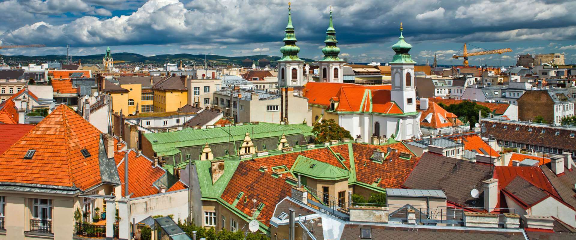 Luftaufnahme von Wien, Österreich, mit Terrakotta- und grünen Dächern, Kirchtürmen mit grünen Kuppeln und einem bewölkten Himmel.
